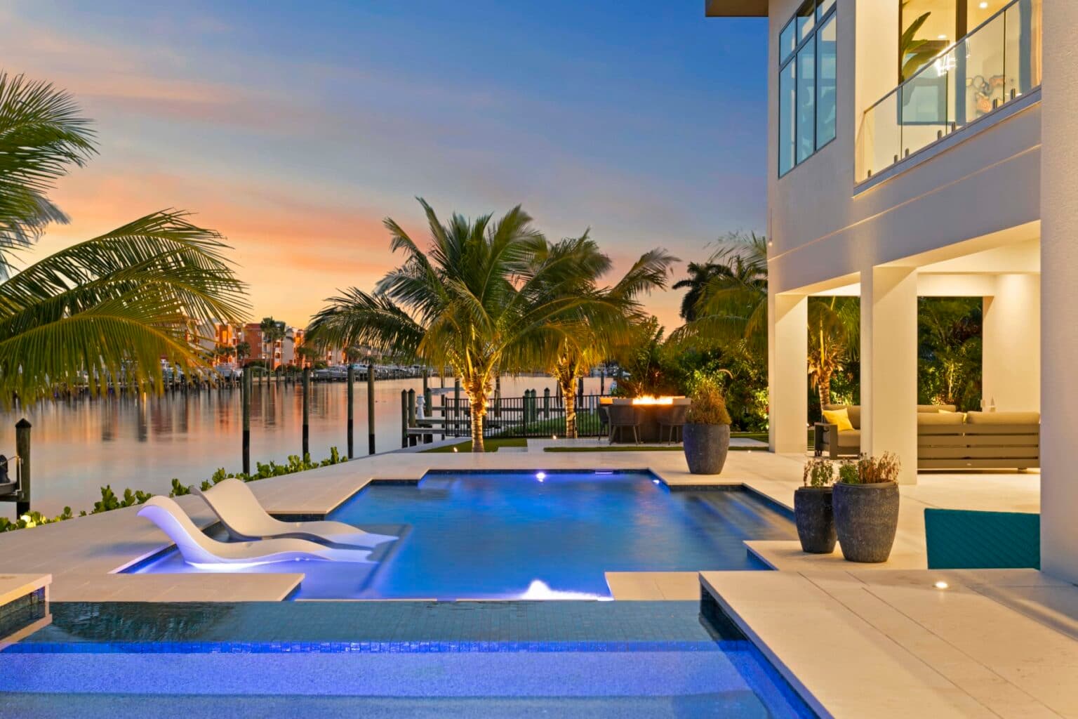 Illuminated pool and loungers beside a waterfront home at sunset